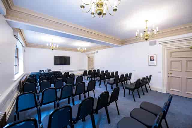 John Tudor presenting in a sophisticated seminar room with black chairs and large screen.