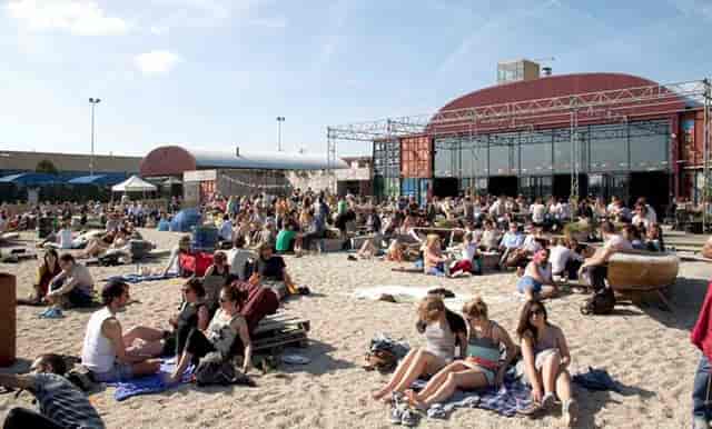 Vibrant outdoor event at Pllek in NDSM with modern architecture and sandy seating.