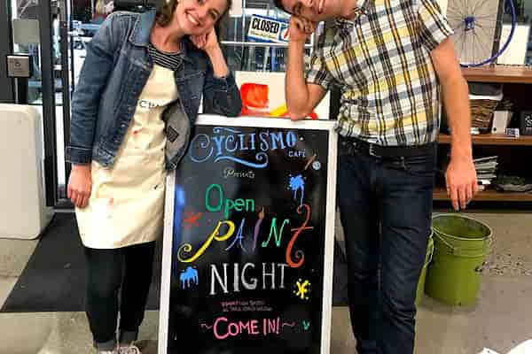 "Open Paint Night sign at Cyclismo Cafe with cheerful hosts and vibrant graphics."